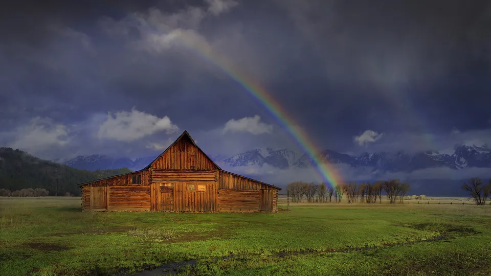 A wooden barn sits in a green field with a rainbow arching across a dark, cloudy sky and mountains in the background.