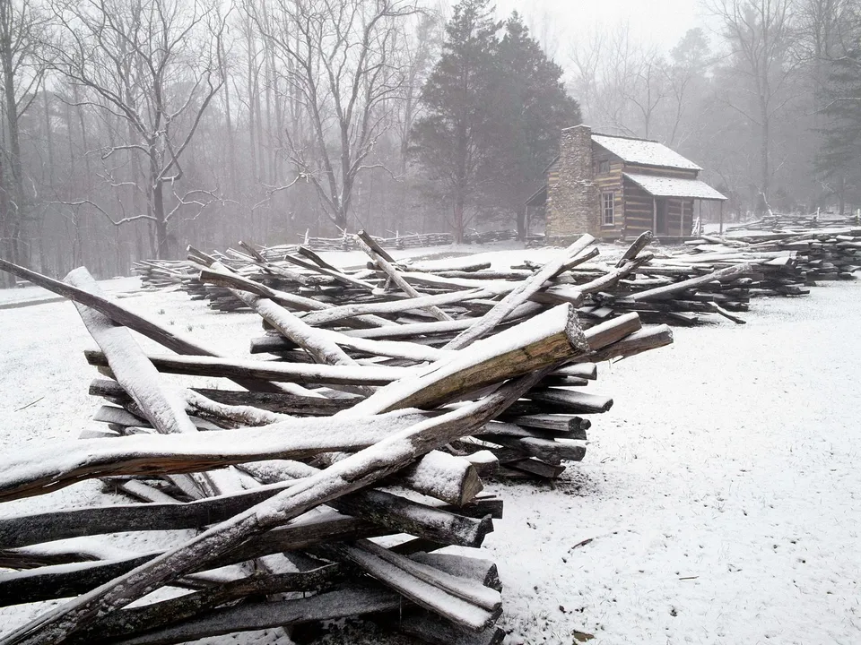 A rustic log cabin with a stone chimney stands in the snow, bordered by wooden split-rail fences.