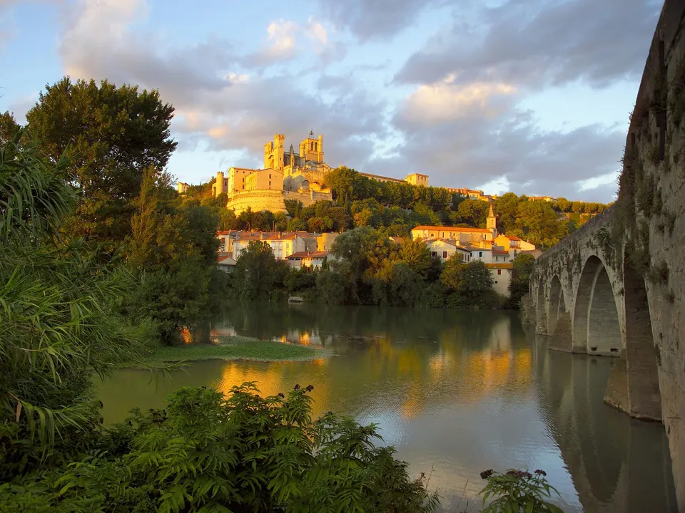 A historic town with a large cathedral sits on a hill overlooking a river, with an old stone bridge on the right.