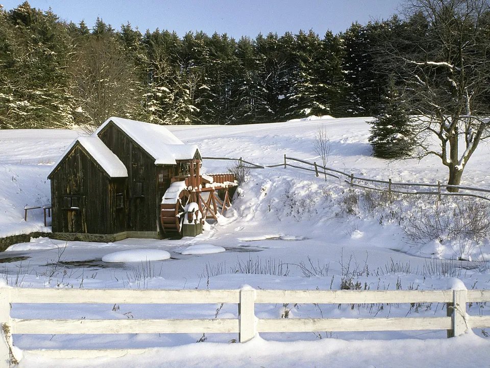 A wooden watermill with a water wheel sits beside a frozen stream, surrounded by snow and pine trees.