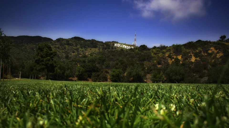 The famous Hollywood sign is visible on a green, hilly landscape under a bright blue sky with scattered clouds.