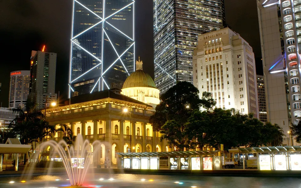 A nighttime cityscape featuring illuminated skyscrapers, a domed building, and a fountain in the foreground.