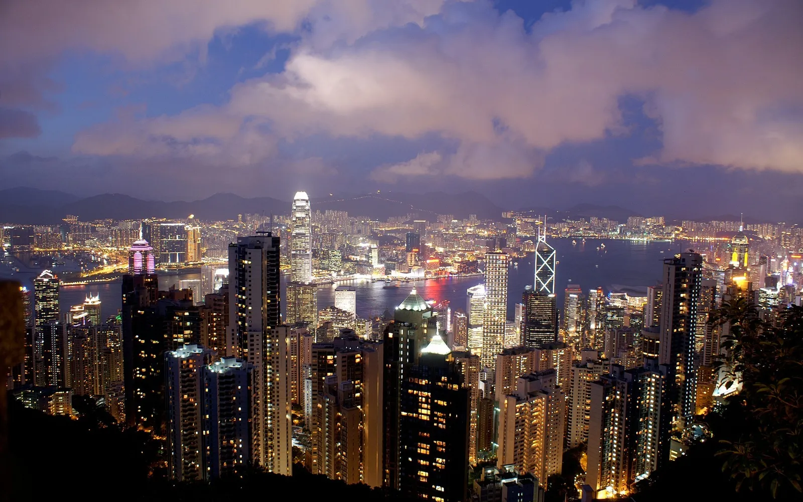 A wide view of the Hong Kong skyline at night, with numerous illuminated skyscrapers and a body of water.