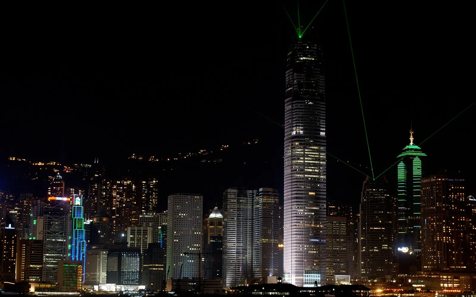 A nighttime cityscape of Hong Kong with illuminated skyscrapers and green laser beams projecting from the top of buildings.