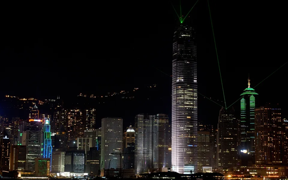 A nighttime cityscape of Hong Kong with illuminated skyscrapers and green laser beams projecting from the top of buildings.
