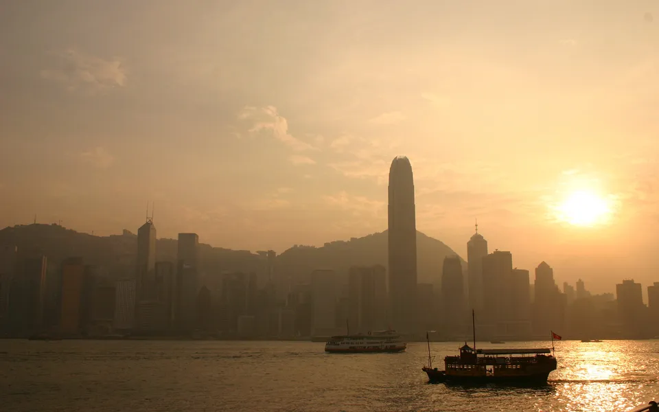 A hazy sunset over the Hong Kong skyline with skyscrapers and boats on the water.
