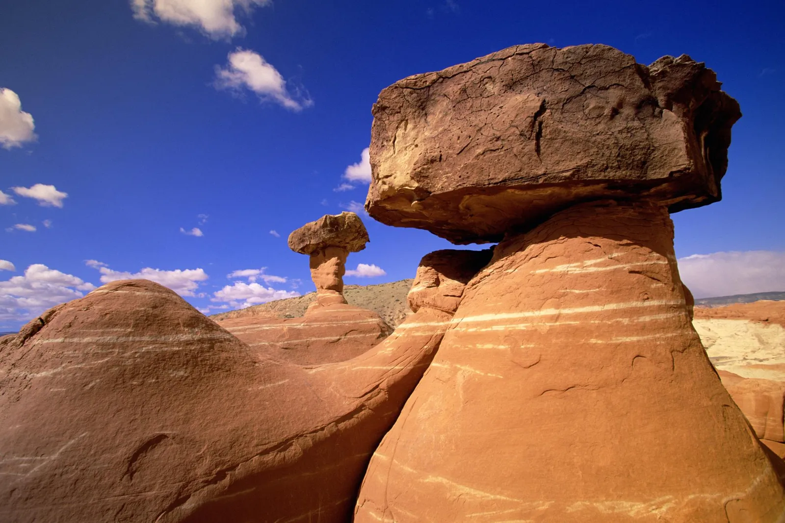 Two large hoodoo rock formations, one with a balanced caprock, stand against a bright blue sky with scattered white clouds.