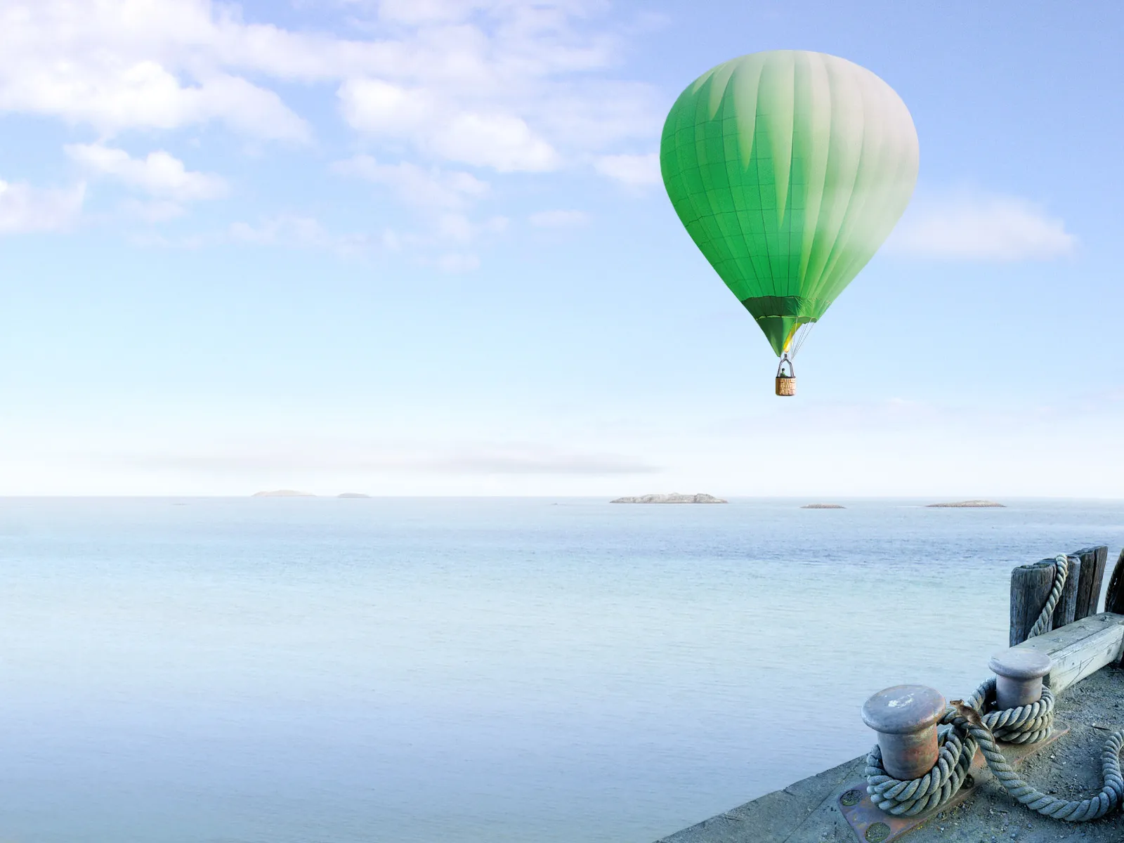 A green hot air balloon floats in a pale blue sky over a calm ocean with distant islands.