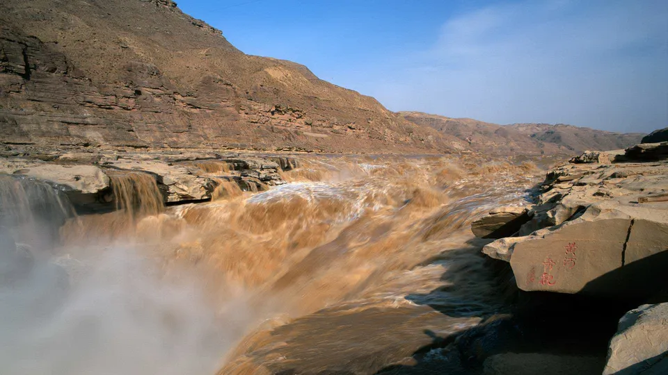 A wide, muddy waterfall cascades over rocky ledges in a dry, mountainous landscape under a clear blue sky.