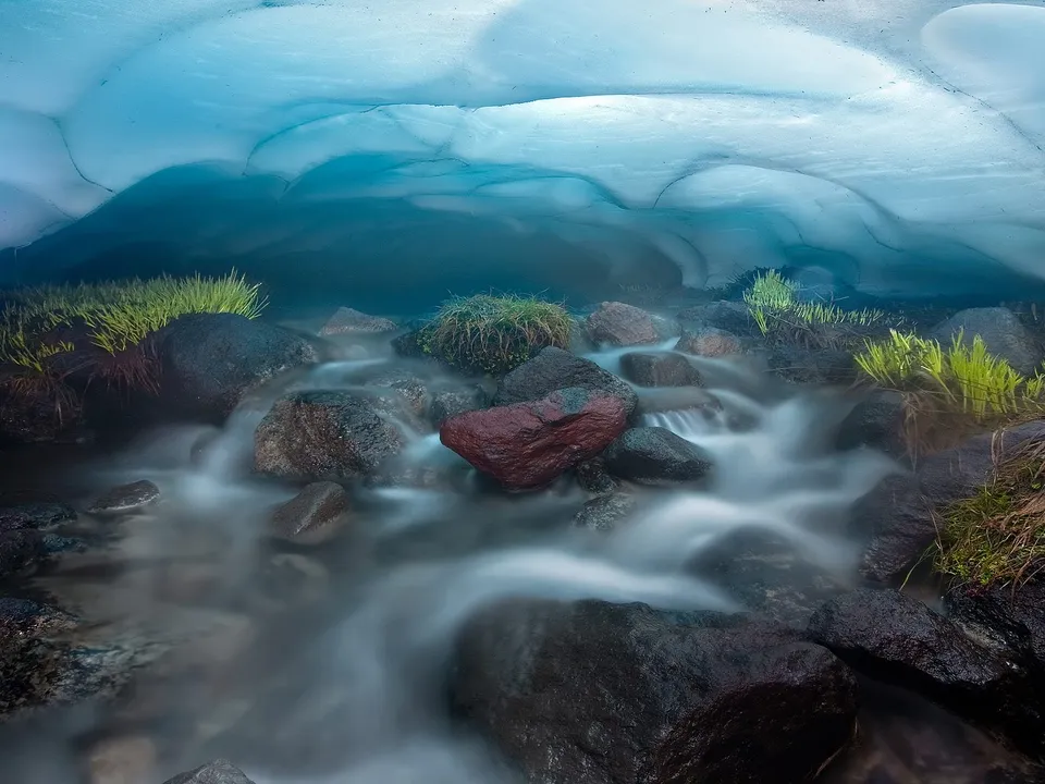 A stream of water flows over rocks inside a glacial ice cave with blue ice overhead.