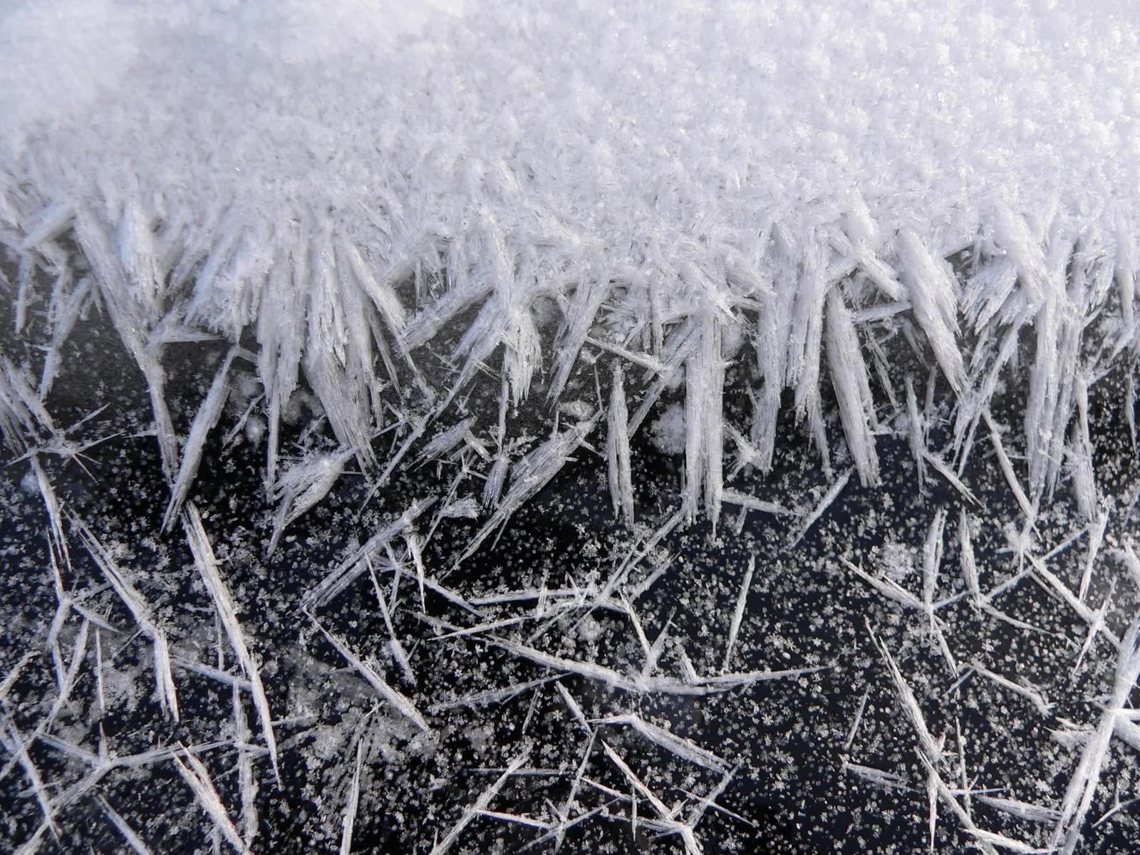 Close-up of sharp, needle-like ice crystals and snowflakes forming on a dark, frozen surface.