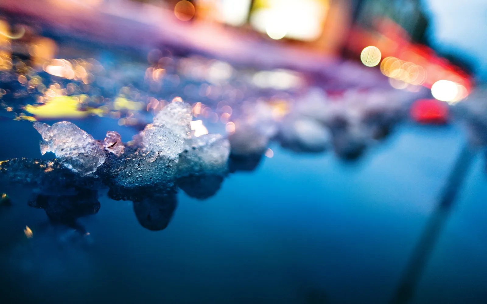 Close-up of glistening ice formations on a dark blue surface, with blurred colorful lights in the background.