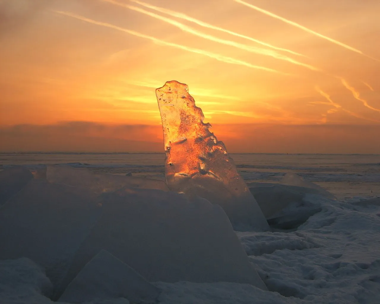 A shard of ice glows orange in the sunset light, with snow-covered ground and a hazy sky.