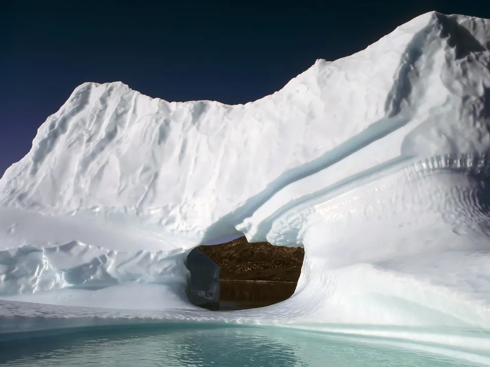 A large iceberg with a natural archway showing dark land and water within, under a dark sky.