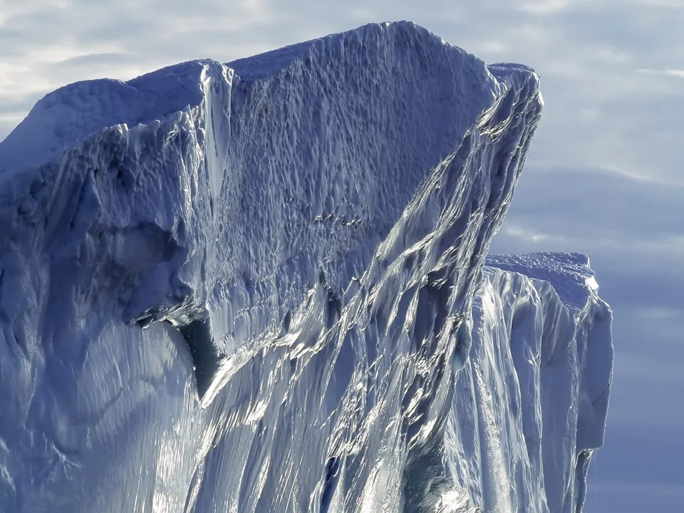 A large, textured iceberg with dramatic light and shadow, set against a cloudy sky.