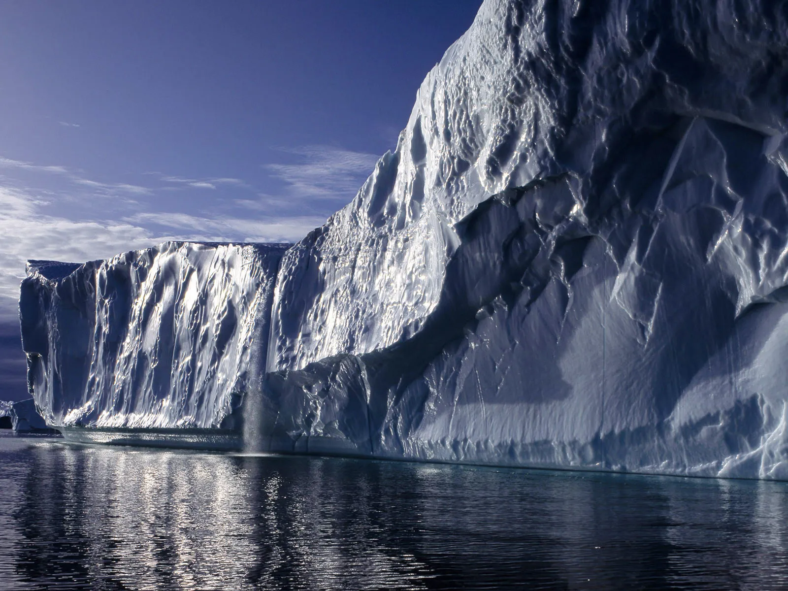 A massive iceberg with a waterfall cascading down its side into the dark ocean under a blue sky.