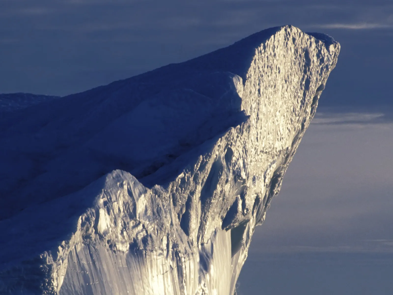 A close-up view of a dramatic iceberg cliff face, illuminated by sunlight against a dark blue sky.