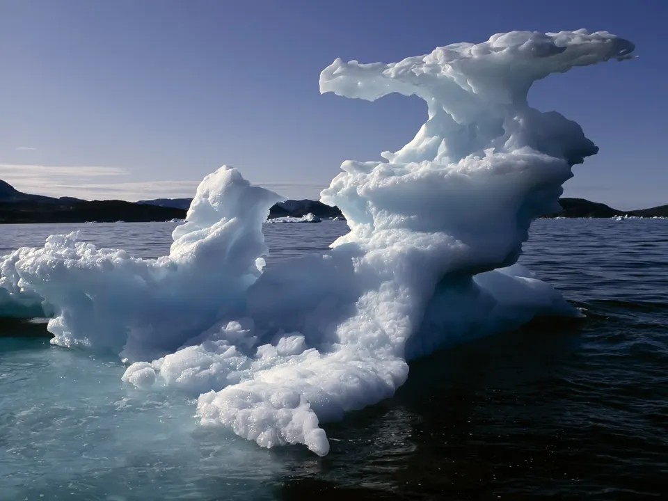 A large, intricately shaped iceberg floats in dark blue ocean water under a clear sky.