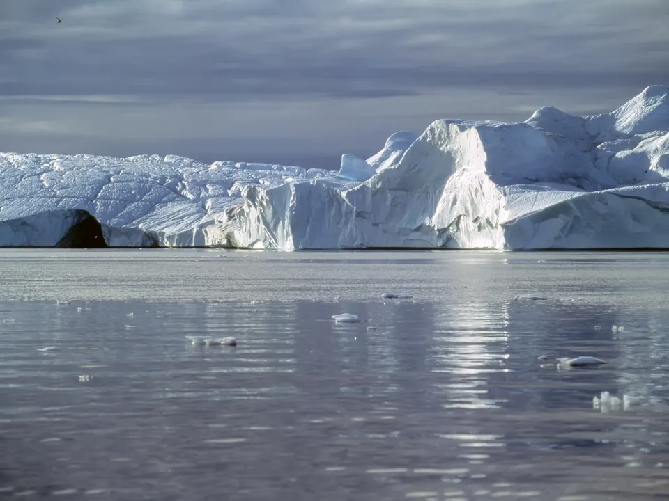 A massive iceberg's textured surface reflects in still water under a dramatic, cloudy sky.