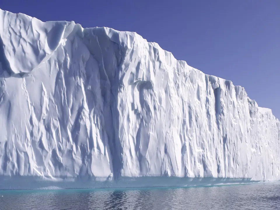 A massive, textured iceberg wall rises from the ocean under a clear blue sky.