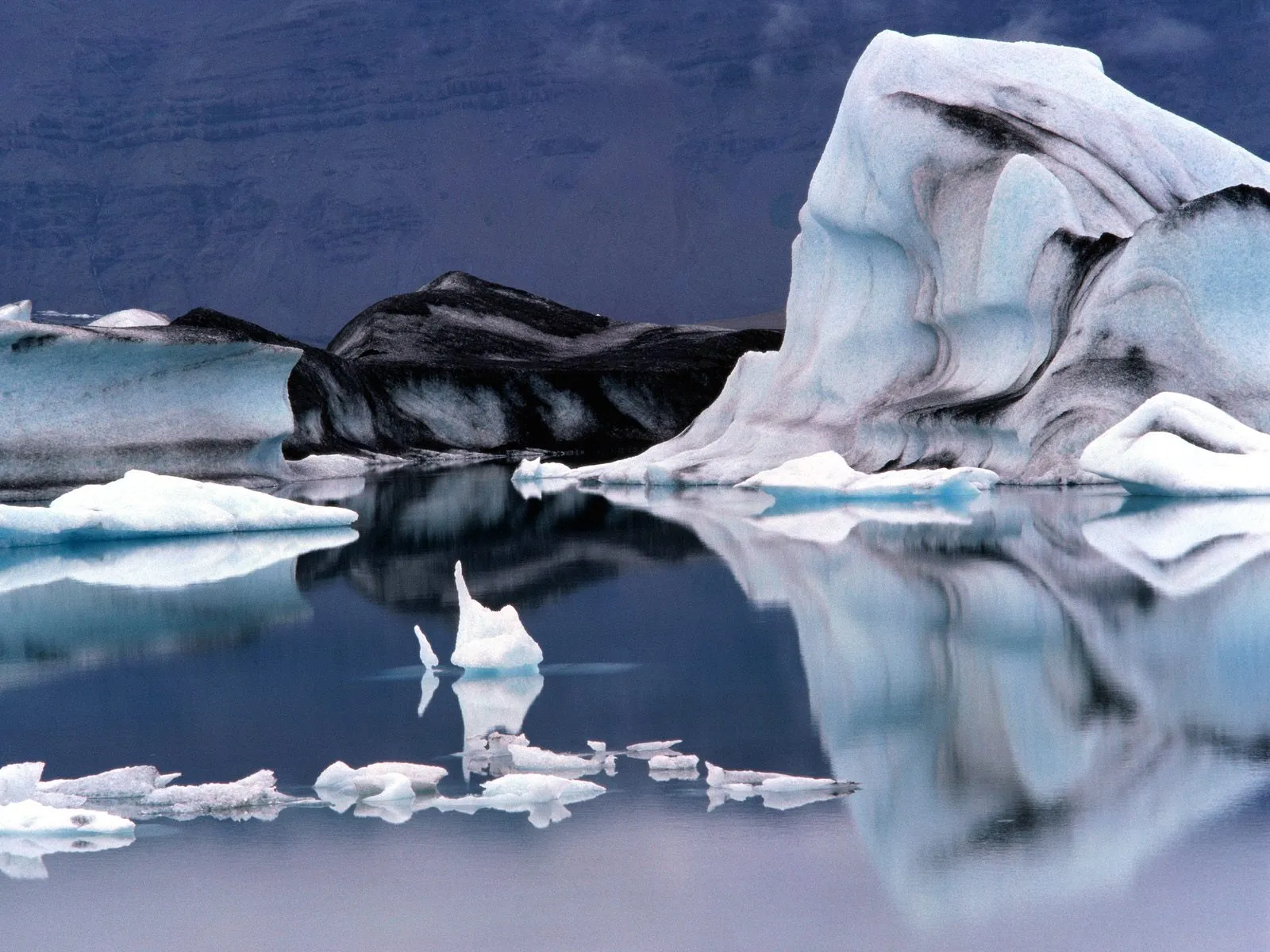 Large icebergs with dark streaks float in a dark blue glacial lagoon, reflecting in the still water.