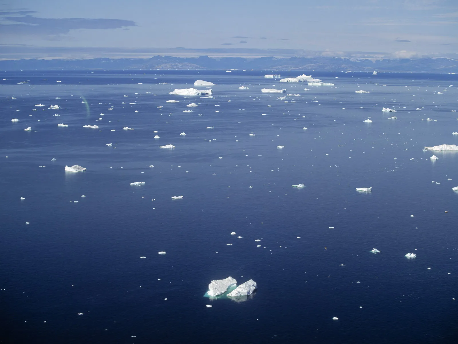An aerial view of a vast, deep blue ocean dotted with numerous icebergs of various sizes under a partly cloudy sky.