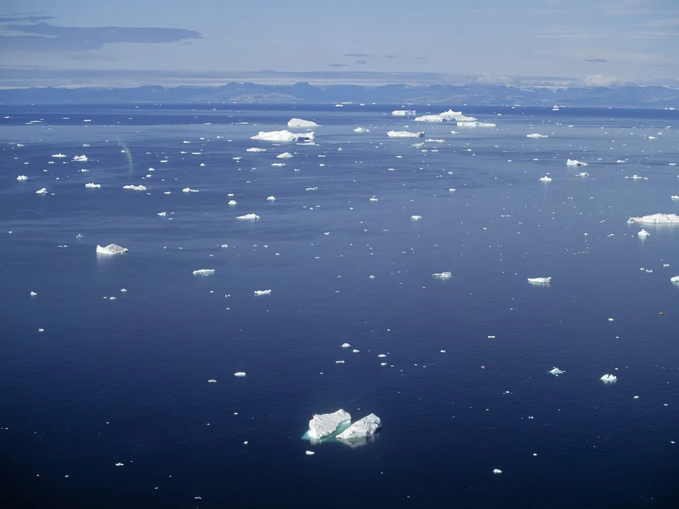 An aerial view of a vast, deep blue ocean dotted with numerous icebergs of various sizes under a partly cloudy sky.