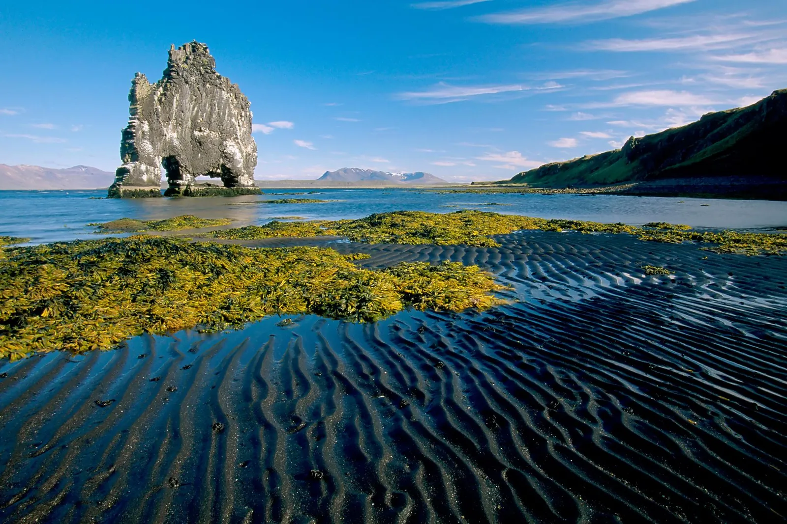 A large rock formation stands in the ocean near a black sand beach with rippled sand and seaweed.