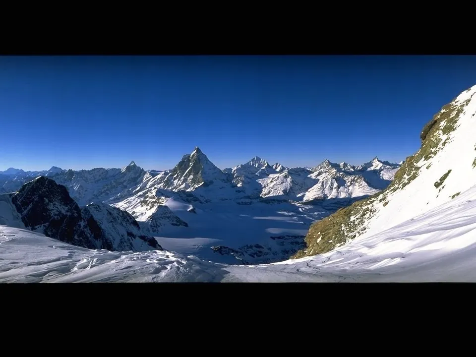 A prominent, sharp mountain peak stands out in a vast, snow-covered alpine range under a clear blue sky.