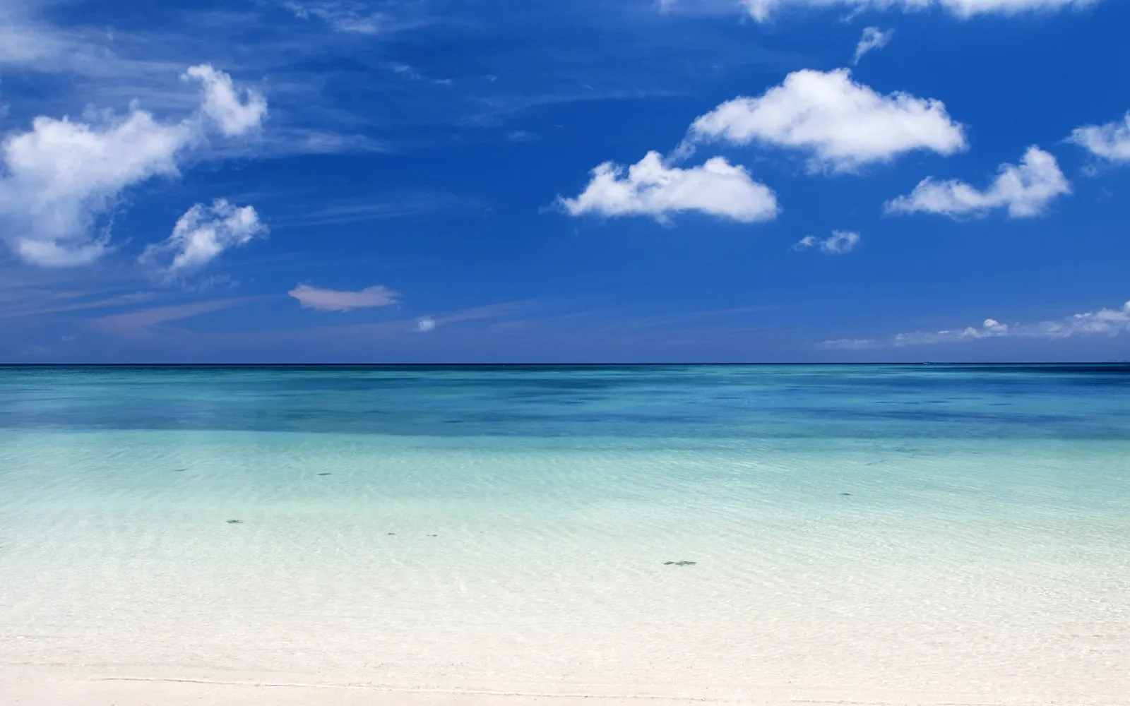 A serene white sand beach with clear, shallow water lapping onto the shore under a blue sky with clouds.