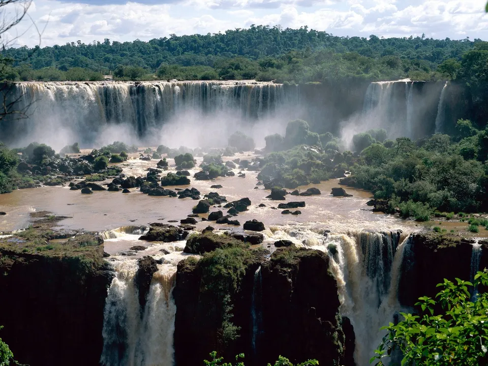 A wide view of the Iguazu Falls with multiple cascades and mist rising, surrounded by dense green forest.