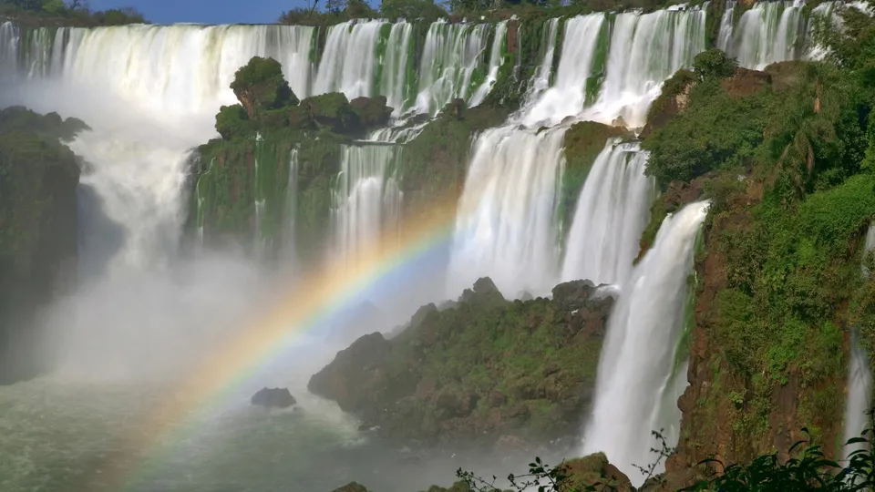 A wide view of Iguazu Falls with a bright rainbow arcing through the mist.