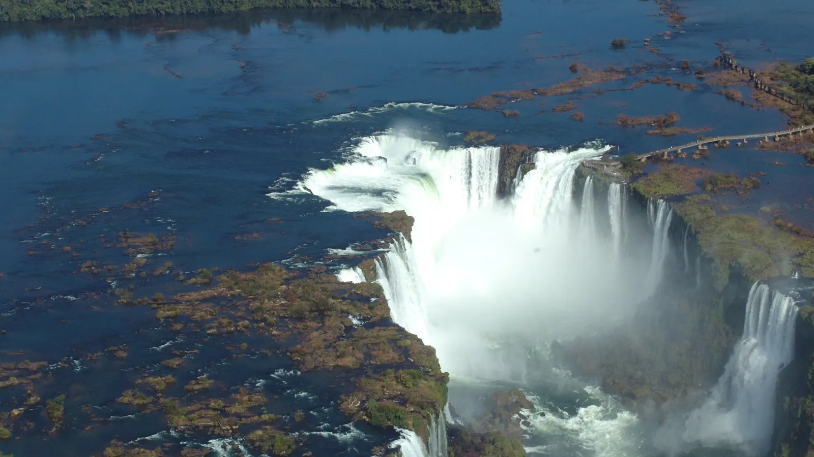 An aerial view of a wide waterfall with lush green vegetation on the cliffs and a bridge over the river.