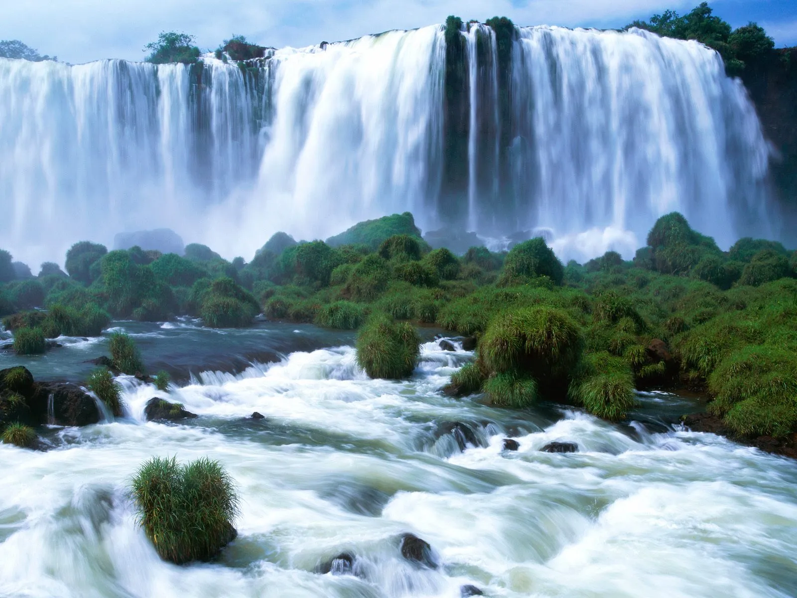 An immense waterfall plunges over a wide cliff with lush green vegetation on rocky outcrops.