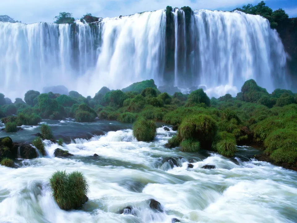 An immense waterfall plunges over a wide cliff with lush green vegetation on rocky outcrops.