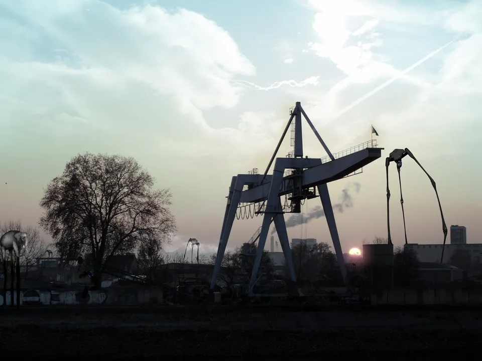 Large industrial cranes stand silhouetted against a pastel sky with a setting sun.