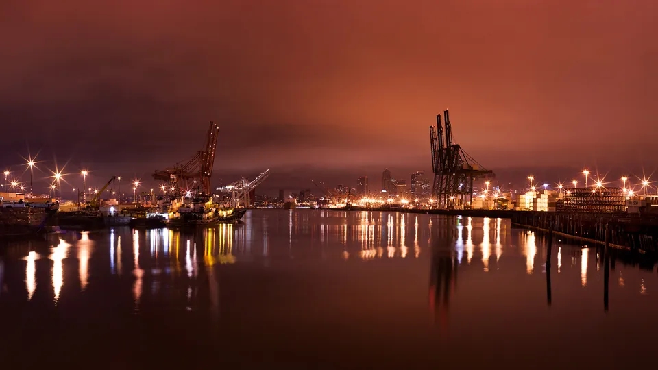 A nighttime view of a busy industrial harbor with large cranes and ships, illuminated by numerous lights reflecting on the water.