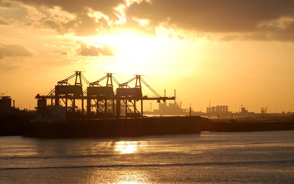 Silhouettes of cranes and ships against a bright, cloudy sunset over a body of water.