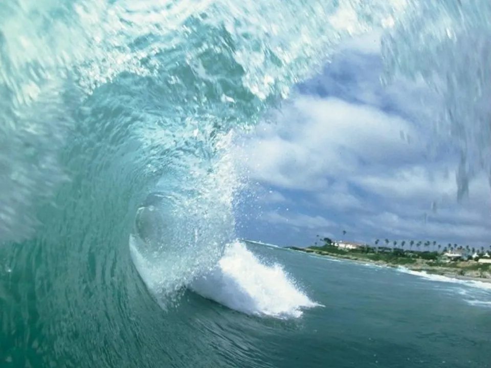 View from inside a breaking ocean wave, looking out towards a cloudy sky and a distant coastline with buildings and palm trees.