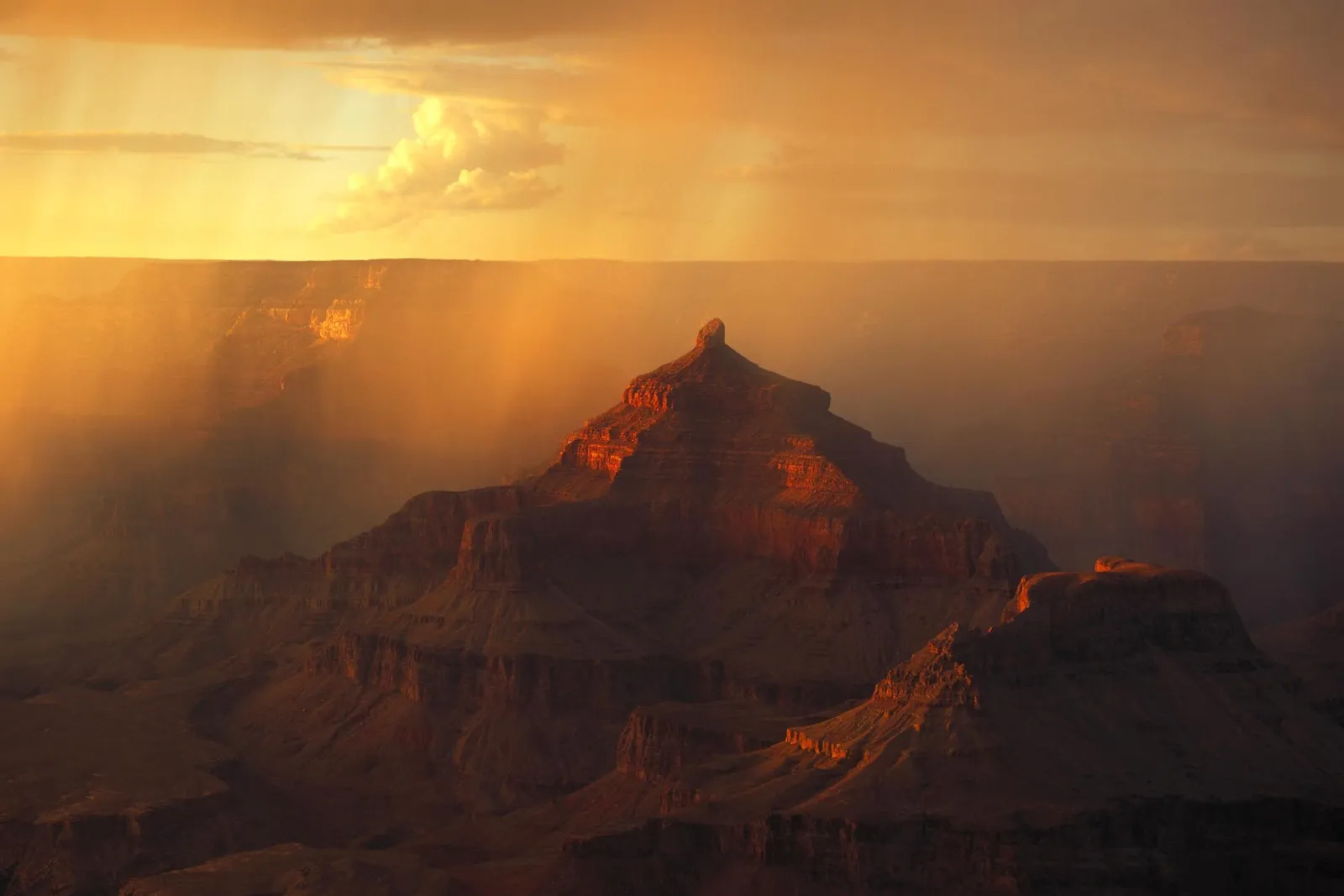 Sunbeams stream through hazy air onto the layered rock formations of the Grand Canyon at sunset.