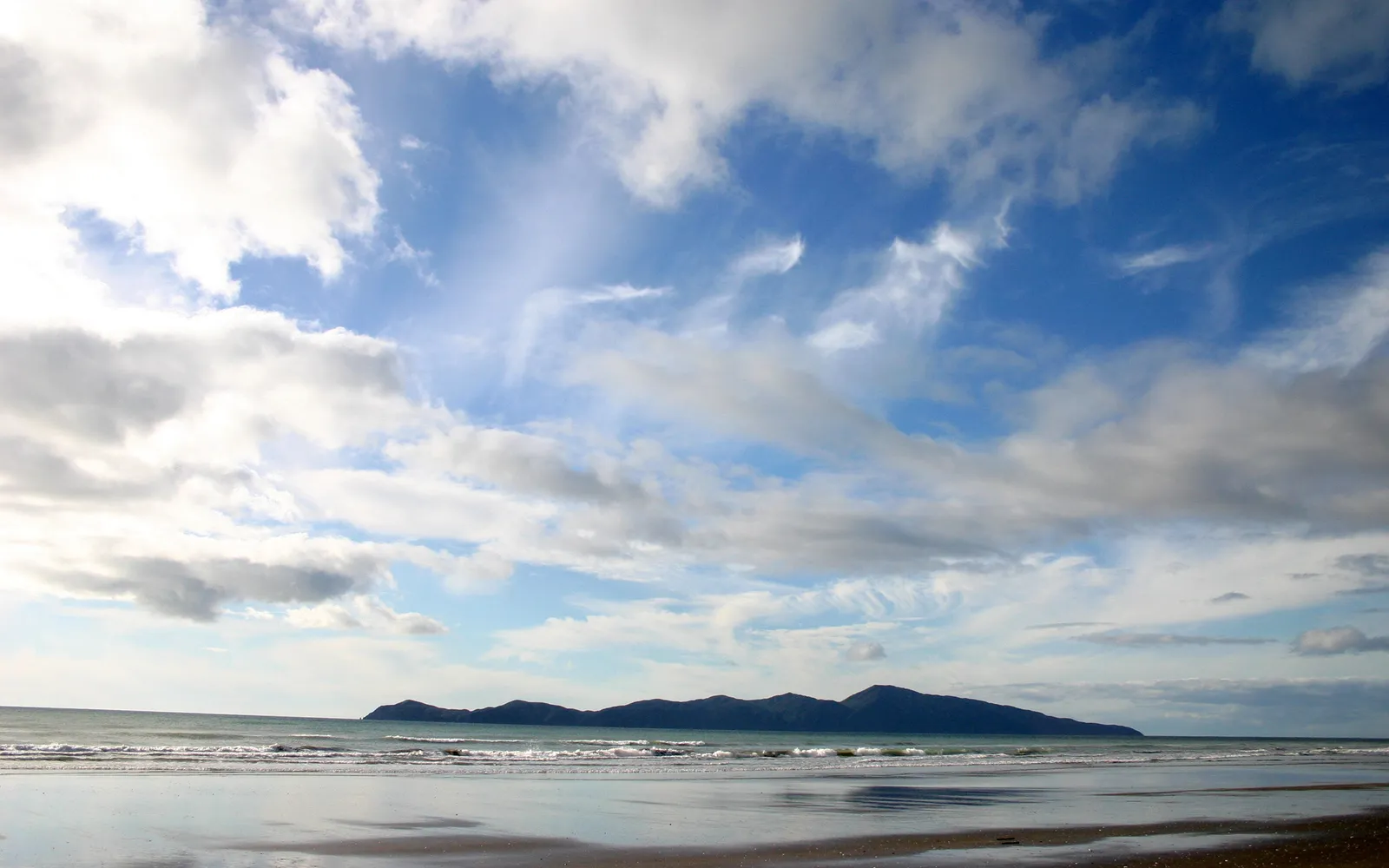 A distant island is visible across the ocean from a wet sandy beach with clouds in the sky.