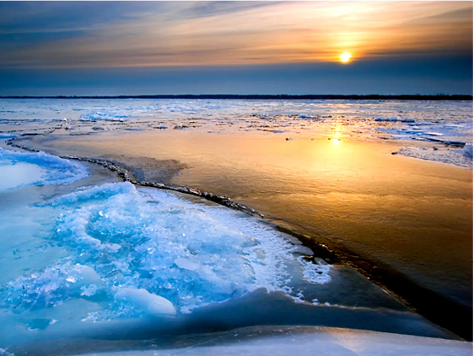 Jagged ice formations in the foreground contrast with a partially frozen lake reflecting a golden sunset.
