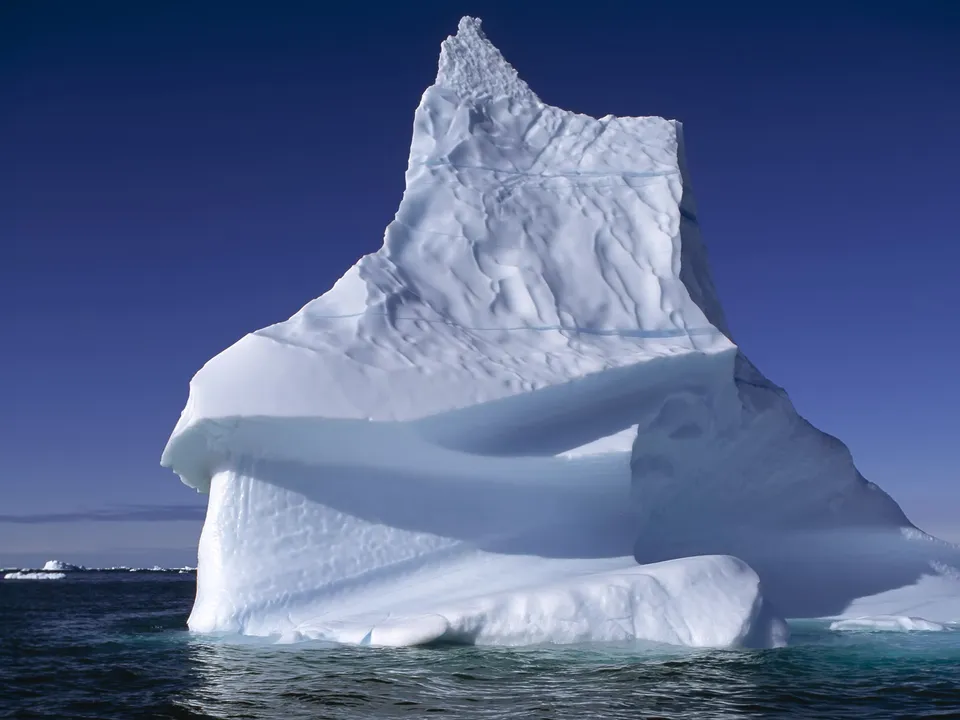 A tall, jagged iceberg stands prominently against a deep blue sky.