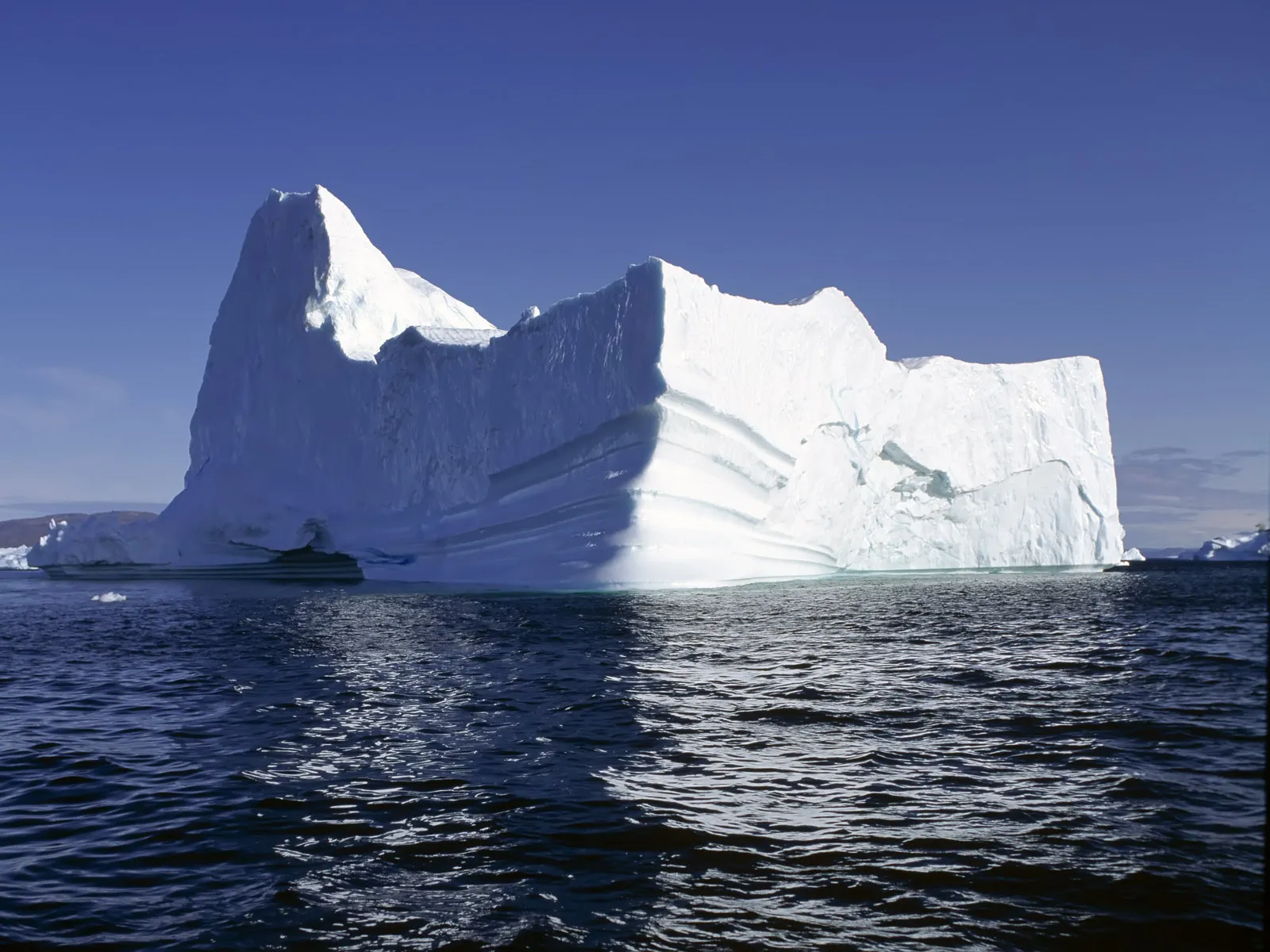 A tall, jagged iceberg is illuminated by bright sunlight, casting shadows on the deep blue ocean.