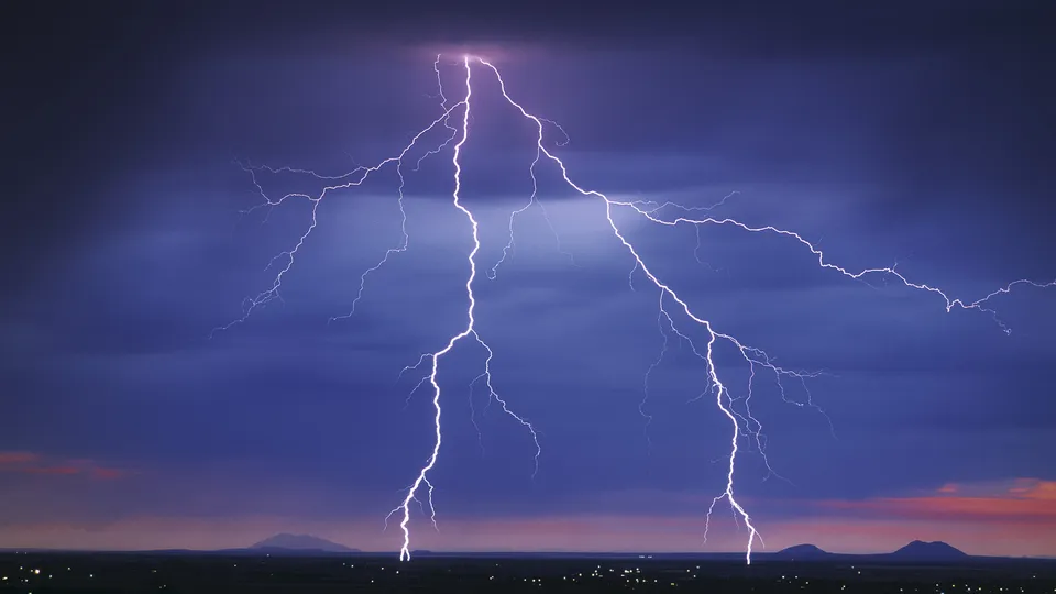 Multiple lightning strikes illuminate a dark, stormy sky at twilight with faint lights from a distant settlement visible.