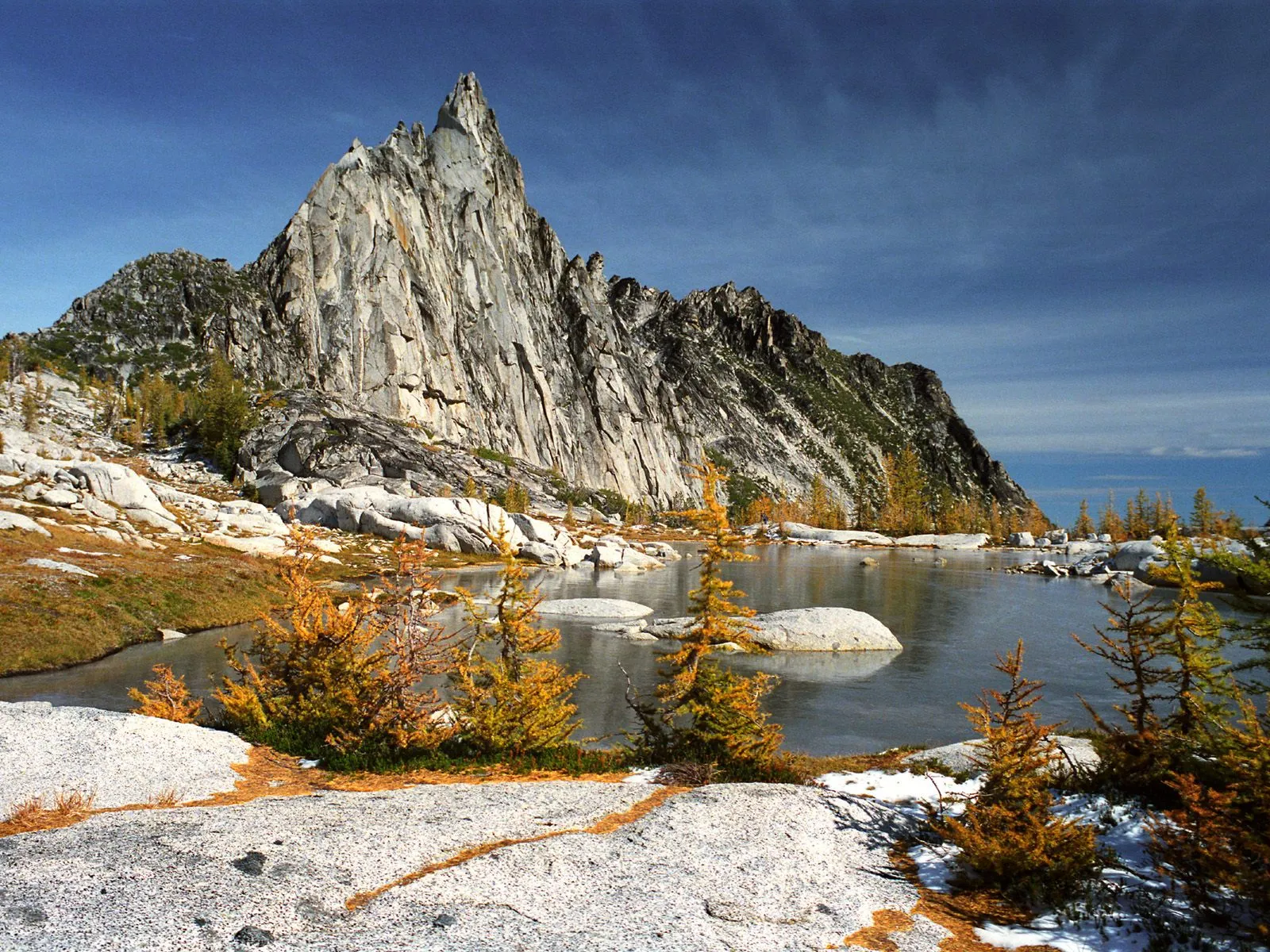 A sharp, rocky mountain peak rises above a calm lake with autumn foliage and patches of snow.