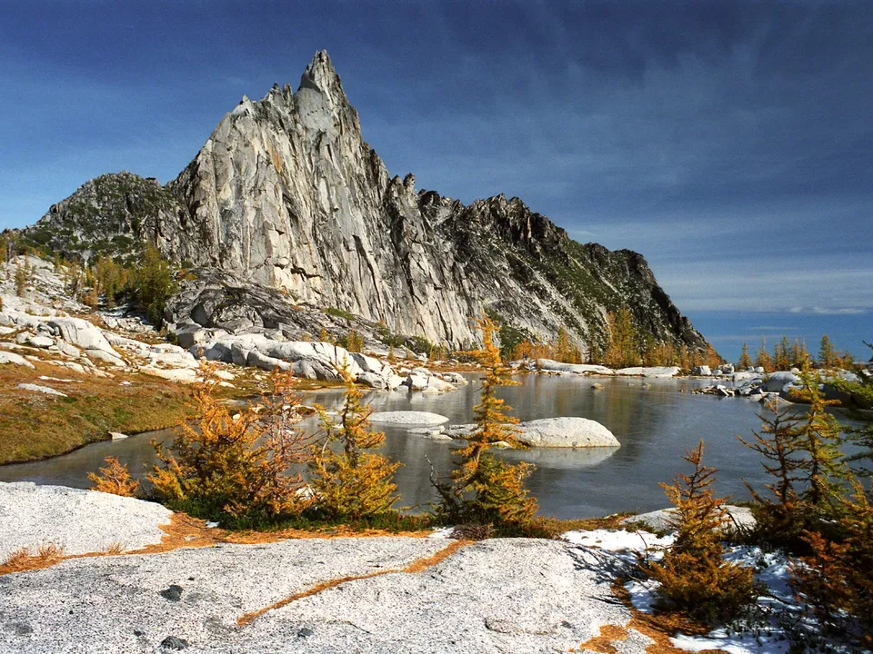 A sharp, rocky mountain peak rises above a calm lake with autumn foliage and patches of snow.
