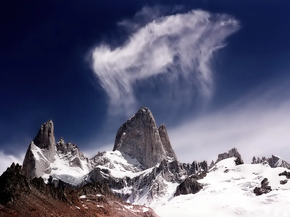 Dramatic jagged mountain peaks covered in snow, with a wispy cloud formation against a deep blue sky.
