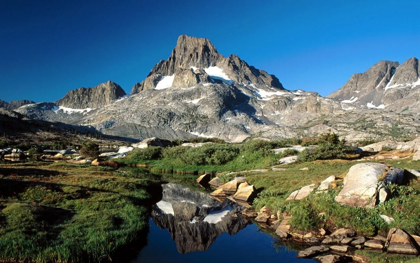 A dramatic, jagged mountain peak under a clear blue sky, reflected in a calm body of water.