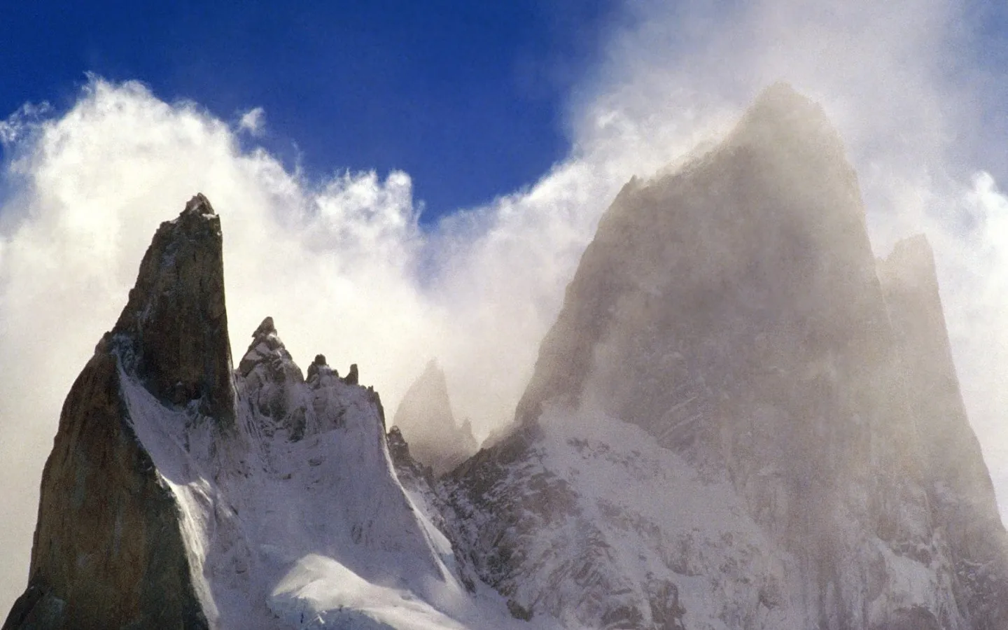 Sharp, snow-dusted mountain peaks pierce through swirling clouds under a bright blue sky.